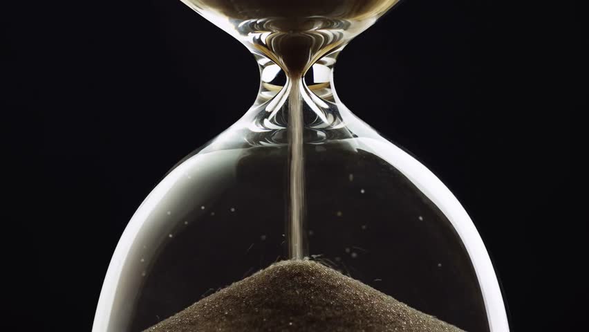 Last seconds flowing in hourglass standing at evening shadow close up. Gold sand running inside transparent clock measuring time indoors. glass egg timer showing countdown on table surface. sandglass