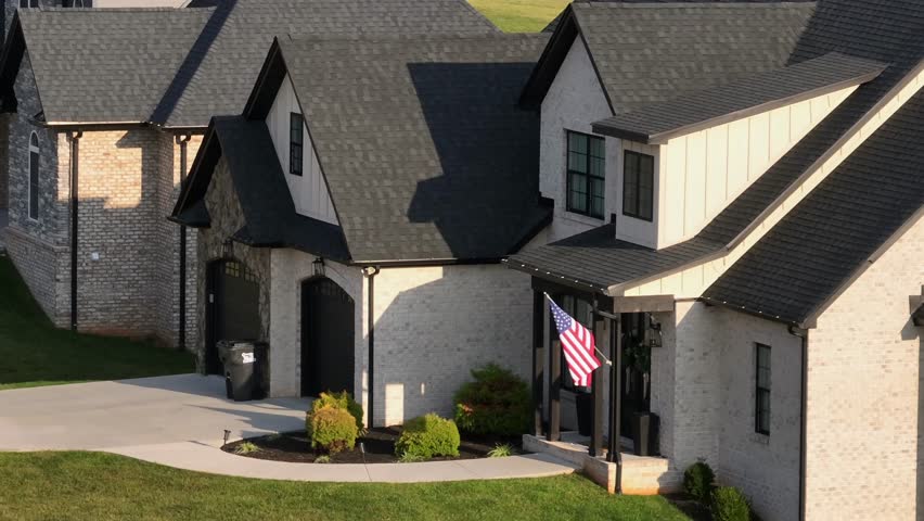 Aerial view of modern suburban home with gabled black roofs on sunny day. Proud american flag waving in front, showing proud and pride community neighborhood in USA. Orbit shot.