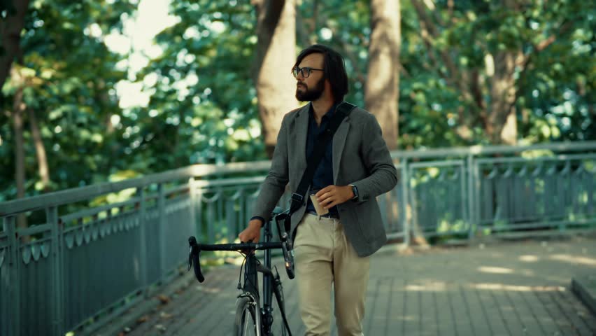 Businessman In Blazer And Glasses Walking With Bicycle In Green Park. Modern Urban Professional Commuting To Work. Freelancer Taking A Break Outdoors. Daily Commute, Sustainable Transportation