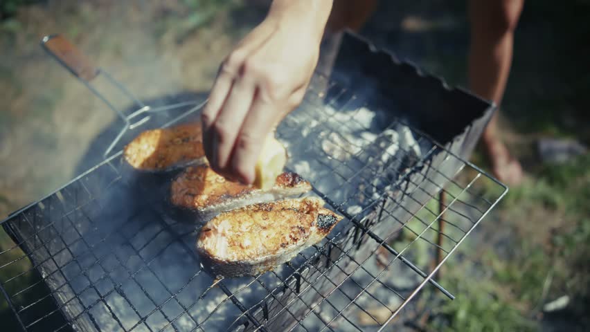 Squeezing Fresh Lemon Juice Over Salmon Steaks Grilling On Barbecue Rack Over Charcoal Fire. Preparing Healthy Outdoor Meal, Delicious Red Fish Cooking On BBQ, Summer Backyard Party And Weekend Picnic