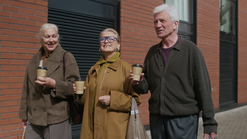 Tracking shot of group of three senior Caucasian friends walking along street, holding shopping bags and coffee cups, enjoying their weekend together