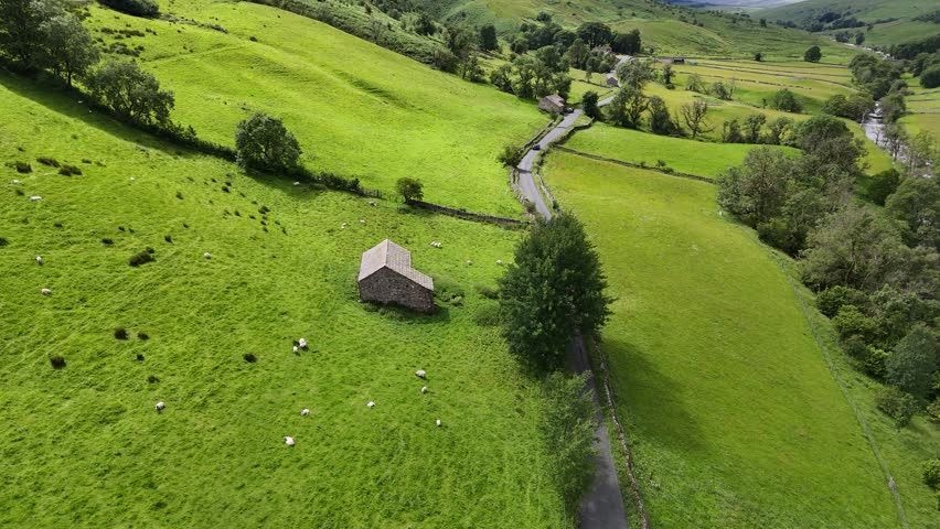 Aerial view of a stone barn nestled amid vibrant green fields, alongside a winding road that cuts through the landscape, Hawes, England, United Kingdom.