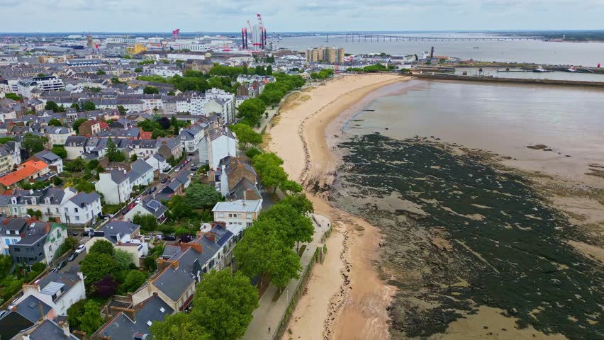 Petit Traict beach in Saint-Nazaire, cityscape, port, and Saint-Nazaire Bridge over Loire estuary in background, France. Aerial forward