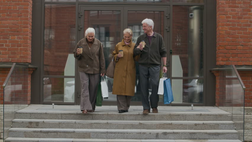 Group of three cheerful senior friends going out of building with coffee cups and shopping bags on autumn day