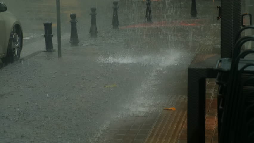 Heavy rain falling on city street from building roof creating puddles. Torrential rain pours, splashing against bollards, with parked cars visible in background during gray, overcast summer day.