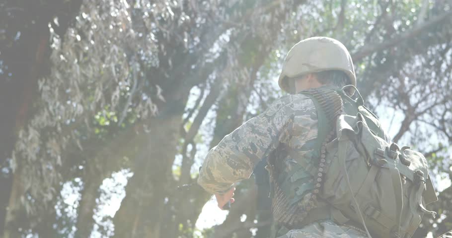 Soldier emerging from foliage in sunlit forest gripping rifle scanning with defense data overlays. Military, tactical, surveillance, technology, forest, security, action
