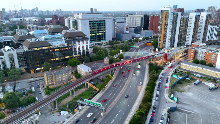 Aerial view of Docklands Light Railway train crossing above a busy highway at dusk, surrounded by modern buildings and city traffic in East London.