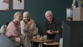 Three senior male and female friends having fun watching TV show and discussing it while sitting in living room - Powered by Shutterstock - Get 15% off with code: PIKWIZARD15