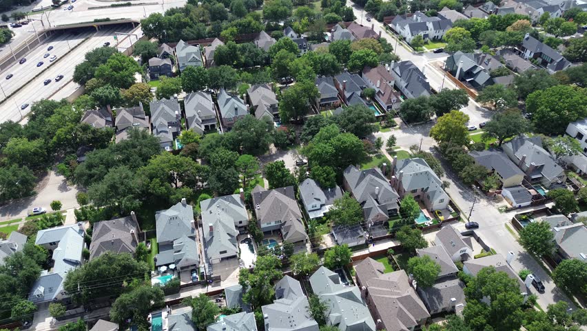 Affluent Highland Park Dallas neighborhood with pitched-roof homes, tree-lined streets, backyard pools, organized grid layout, located adjacent to US 75 highway with visible traffic and overpass. USA