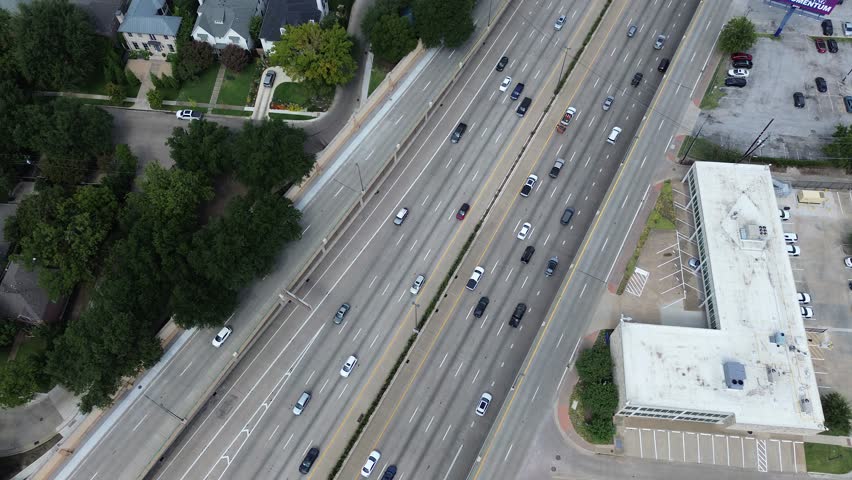 Dense traffic flow along US 75 in Dallas, Texas, multi-lane highway, adjacent tree-lined residential homes on one side, and large commercial building with parking lot on the other, flyover. USA