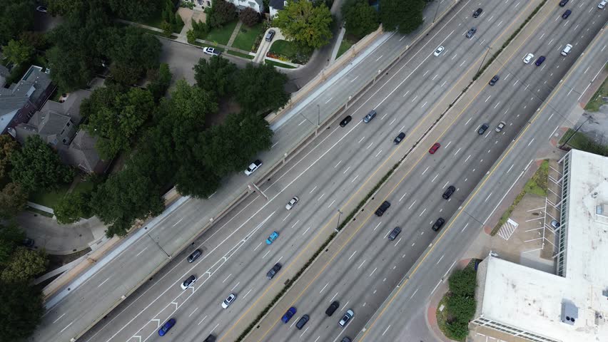 Busy US 75 in Dallas, Texas with multi-lane highway and dense traffic flow, adjacent tree-lined residential homes on one side, large commercial office building with parking lot on the other. USA