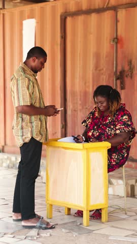 Vertical Video, Mobile money (MoMo) agent assisting a customer with a transaction at an outdoor stand. Digital payments, fintech and financial inclusion in Africa.