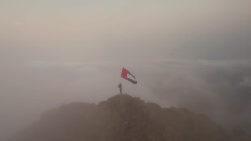 Drone footage of low clouds hugging the peaks of the mountains of Fujairah and Ras Al Khaimah in the UAE as the sun rises in the morning, and a person carrying the UAE flag fluttering in a cinematic 