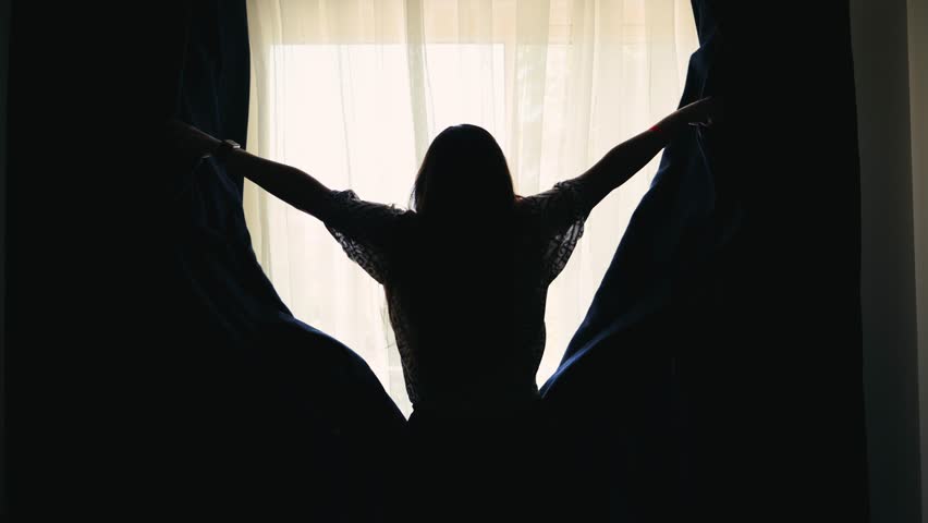 The woman pulls back the curtains and opens the morning light inside.