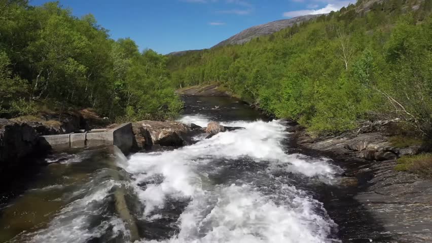 A clear blue sky above a rushing river flowing through a lush green forest and rocky terrain.