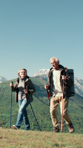 Slow motion vertical video senior couple trekking mountain trail with backpacks and poles healthy lifestyle active holiday adventure travel nature freedom wellness summer outdoors