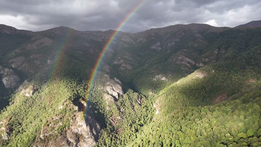 Aerial drone footage of lush forest mountains with a vibrant rainbow under cloudy sky