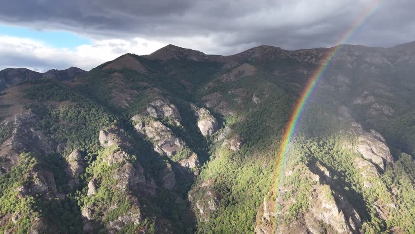 Aerial drone footage of lush forest mountains with a vibrant rainbow under cloudy sky