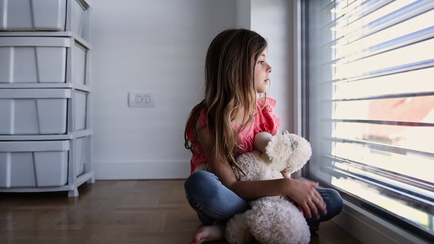 Young girl sits cross-legged on a wooden floor, clutching a teddy bear and gazing through slatted blinds. A quiet, contemplative domestic scene evoking childhood, longing, and solitude.