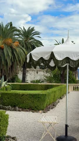 Cinematic view of old-school black-and-white sunshade with white table and chairs in lush garden at Isola del Garda, Italy. Venetian villa, vibrant flowers, elegant outdoor lounge lake view 