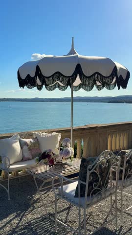 Cinematic view of old-school black-and-white sunshade with white table and chairs in lush garden at Isola del Garda, Italy. Venetian villa, vibrant flowers, elegant outdoor lounge lake view 