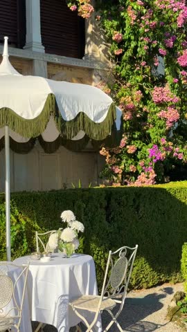 Old-school black-and-white sunshade with white table and chairs in lush garden at Isola del Garda, Italy. Venetian villa with bright floral bushes, elegant outdoor relaxation area
