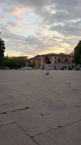Curly-bearded dad riding longboard with toddler daughter in shorts across Verona city square. Laughing, playful father-daughter moment, joyful summer outing, happy Father's Day