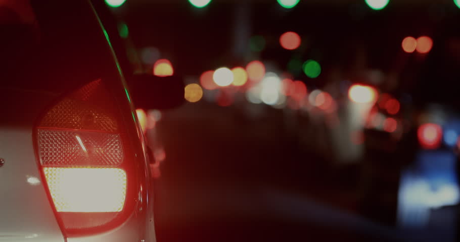 Rear view of car stopped in night traffic, brake light glowing red with stream of blurred headlights and taillights in deep background along crowded road