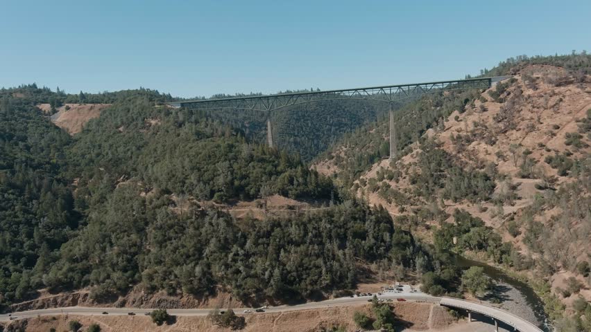 Drone panning shot over hills with winding road, river, and Foresthill Bridge in Sierra Nevada foothills.