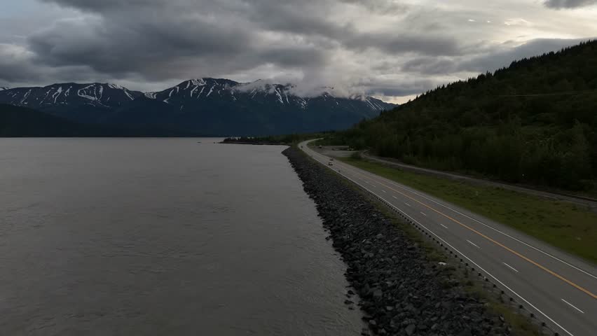 Aerial View of Seward Highway heading North to Anchorage Alaska, Late June, Few cars driving down road, turnagain arm, Black mountains with snow lines, cloudy skies,
