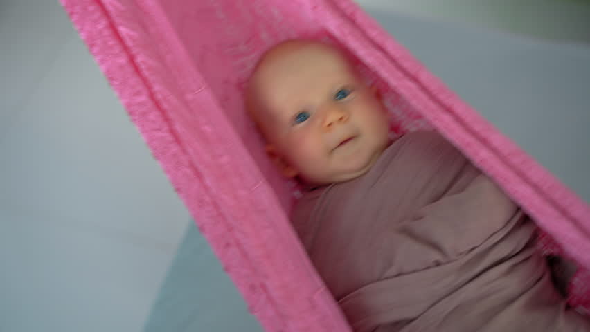 Loving mother gently swings her swaddled two-month-old baby in a hammock to help him fall asleep, creating a peaceful and nurturing family bedtime moment.