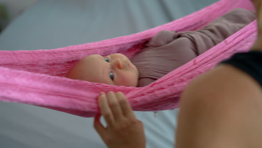 Loving mother gently swings her swaddled two-month-old baby in a hammock to help him fall asleep, creating a peaceful and nurturing family bedtime moment.