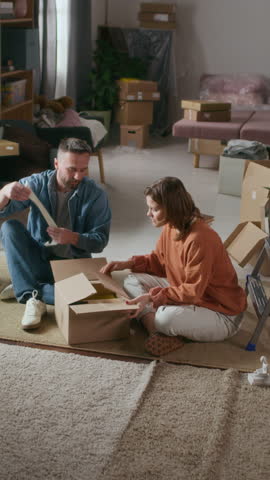 Vertical shot of man and woman unpacking cardboard boxes with their belongings while their little son trying to have fun during their relocation to new home