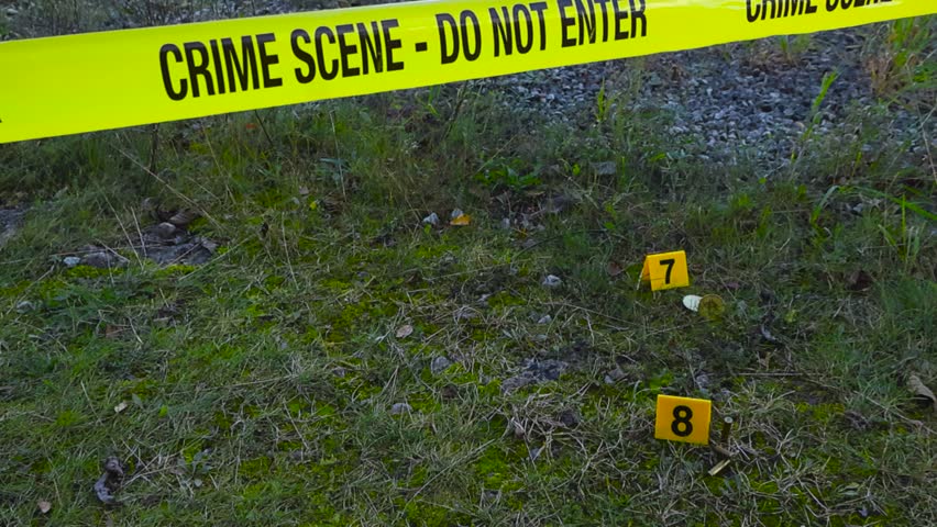 Yellow colored police line invstigation forensic crime scene tape in front of a grassy and gravel area where police crime markers are placed next to bullets and cryptocurrency Bitcoin coins, day time.