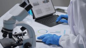 Scientist wearing lab coat and gloves analyzing data and charts on tablet and paper, with microscope and laboratory glassware in background - Powered by Shutterstock - Get 15% off with code: PIKWIZARD15