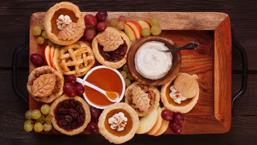 Autumn pie theme dessert board. Top down view stop motion placement video over a dark wood background. Pumpkin, apple and pecan mini pies, with whipped cream, caramel and fruit.
