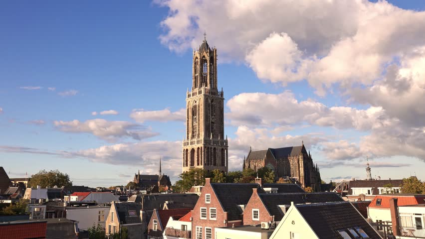 Historic Dom Tower in Utrecht with Gothic architecture rising above city rooftops under blue sky, symbol of Dutch heritage and culture. Clouds move quickly through the timelapse.