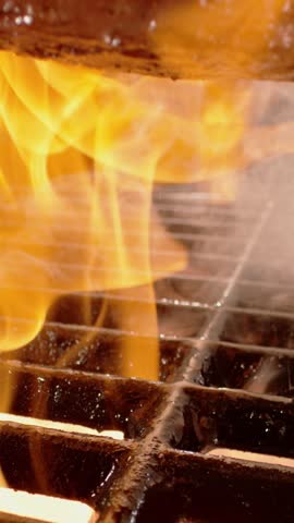 Super slow-motion shot of a thick-cut beef steak falling on flaming grill with fire and smoke in the background
