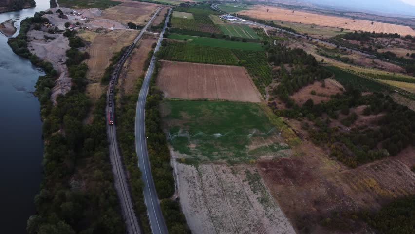 Aerial view of a freight train passing through a rural landscape with a river, fields, road, and trees alongside.