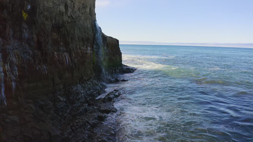 Waterfall cascades from cliff into the Pacific Ocean at Davenport Pier Beach along Highway 1, California, USA. Scenic coastal view with waves, rocky shoreline and blue sky