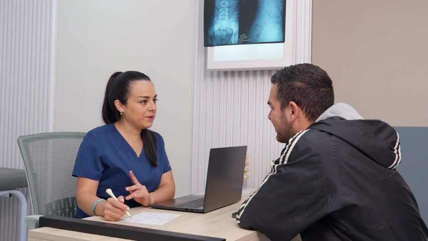 Professional female physiotherapist sitting at her desk, explaining a diagnosis to a male patient with an x ray image in the background