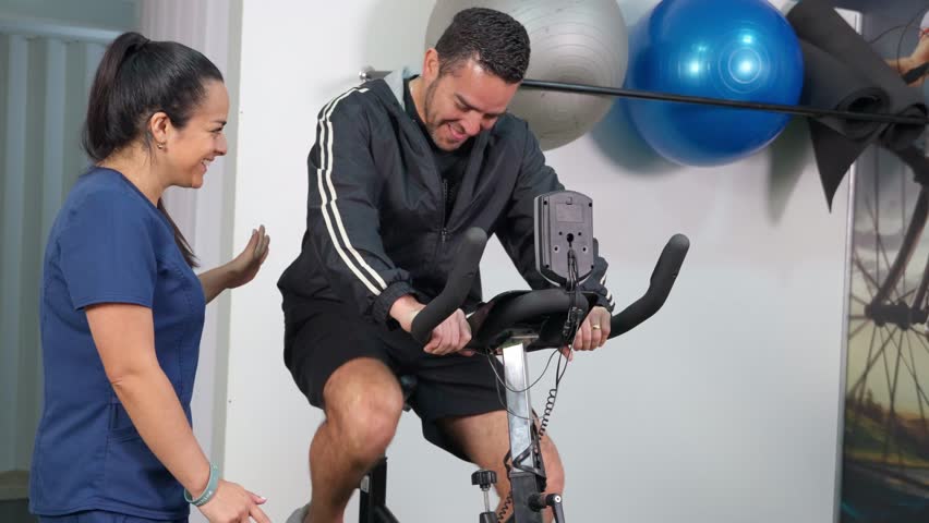 Female therapist guiding a male patient during a rehabilitation session. A man is pedaling on a stationary bike while the doctor assists