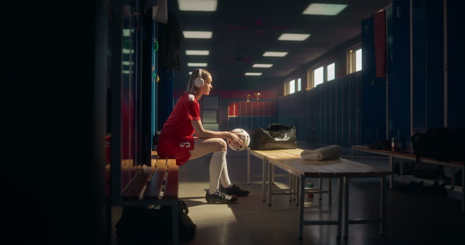 Female Soccer Player in Red Uniform Preparing in Locker Room, Sitting Alone, Listening to Music in Headphones. Athlete is Calm, Preparing Mentally Before Stepping Onto the Field