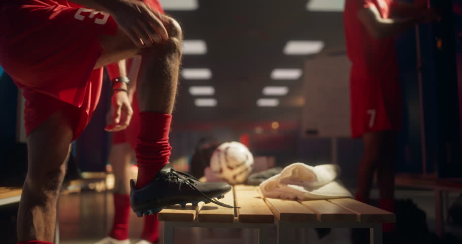 Soccer Athlete in Sports Red Outfit Bending Down to Tighten Laces on His Shoes in a Close Up View. Teammates Around Continue Their Pre-Match Preparation, Getting Ready for a Championship Game
