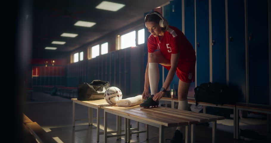 Woman Athlete in Red Soccer Uniform Preparing for a Match a Locker Room, Listening to Music on Headphones as She is Tying Laces on Her Cleats. Female Footballer Gathering Focus Before Training