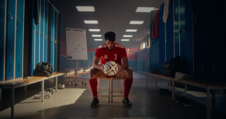 Portrait of a Handsome Young Male Sitting on a Bench in a Locker Room, Playing with a Soccer Ball. Man Wearing a Red Sports Uniform, Looking at Camera with a Confident Smile