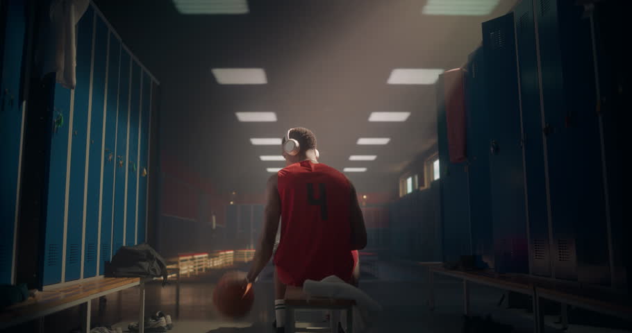 Young Black Athlete on Locker Room Bench, Bouncing the Ball, Spinning It on a Finger and Wearing Headphones. Music Fuels His Concentration and Energy as He Gets Ready for the Match. Back View