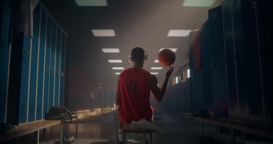 Young Black Athlete on Locker Room Bench, Bouncing the Ball, Spinning It on a Finger and Wearing Headphones. Music Fuels His Concentration and Energy as He Gets Ready for the Match. Back View