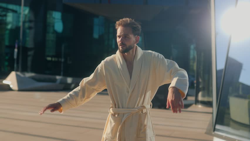 Man in robe and slippers dances freely outside a business center, showing a cheerful and funny side of corporate life against modern reflective skyscraper glass.