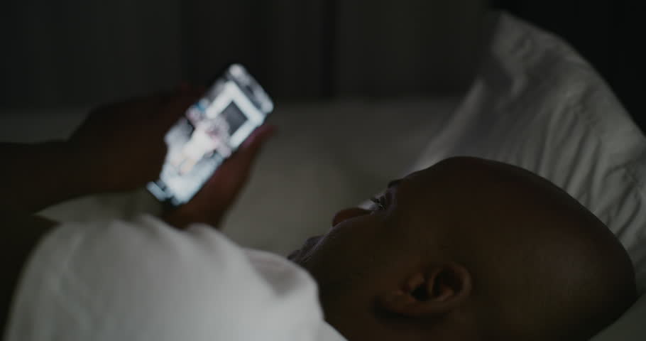 Night, Adult Guy Lies in Bed Under Warm Glow of Bedside Lamp, Scrolling Through Social Media on Smartphone. Concept of Unhealthy Bedtime Habits, Screen Addiction, Poor Rest, and Insomnia. Static Shot.